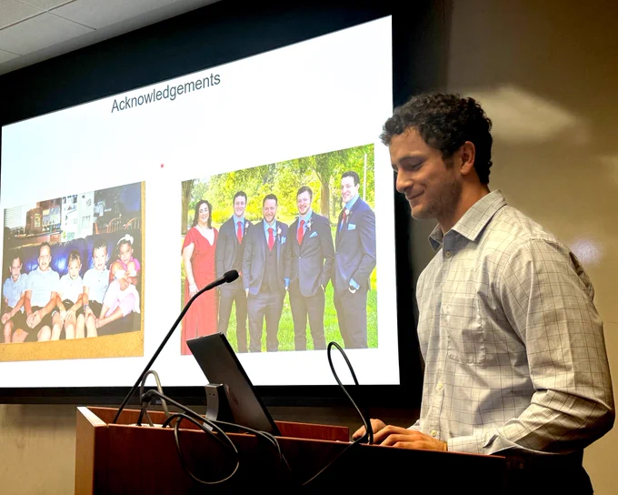 Greg Mannino stands at a lecturn with a mic and his computer, in the background is a projection screen with an Acknowledgements slide showing, which has two personal family pictures displayed.