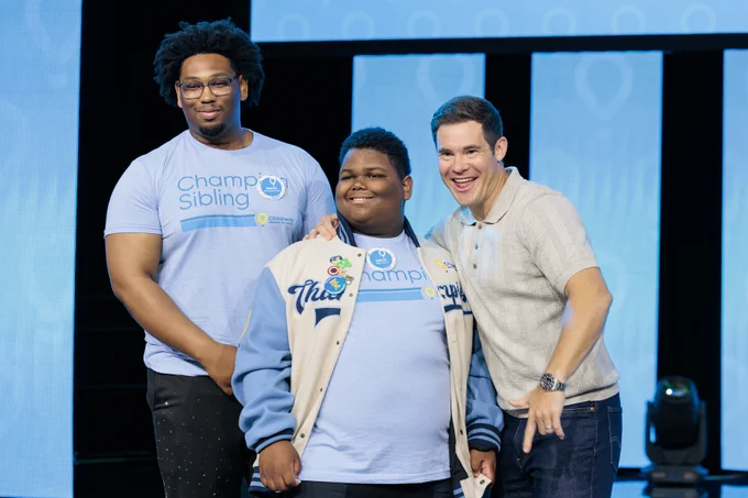 Three individuals stand together on stage, two wearing 'Champ Sibling' shirts, posing for a group photo at an event.