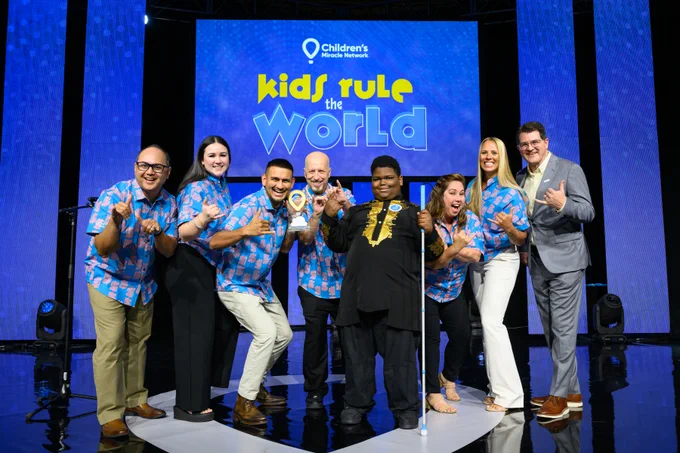 Group of eight people posing on stage with a 'Kids Rule the World' sign in the background at a Children's Miracle Network event.