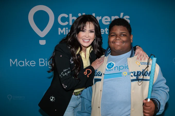 Two individuals posing in front of a Children's Miracle Network backdrop, one wearing a Champion jacket and holding a blue cane.
