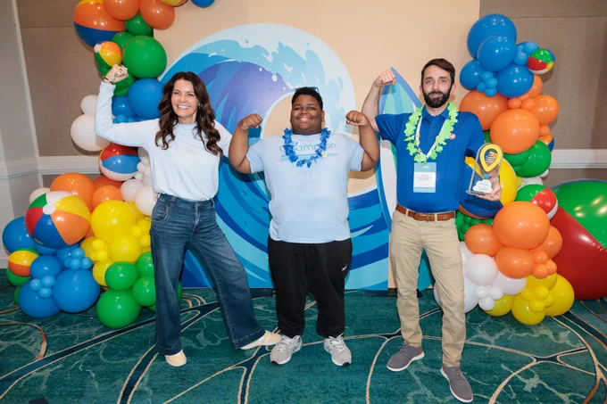 Three people posing with flexed arms in front of a colorful balloon arch and wave-themed backdrop at an event.