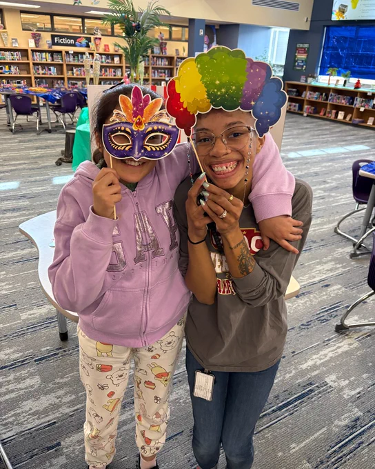 Two people wearing colorful party masks and smiling at the camera in the library.
