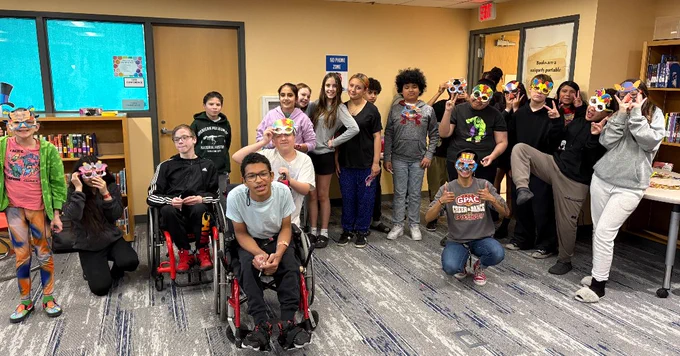A group of people gathered in a classroom setting, some standing, some seated, with smiles on their faces.
