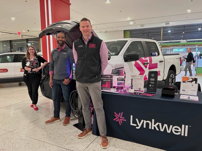Two men stand in front of a white Ford F-150 Lightning electric pickup truck at an indoor exhibition booth for Lynkwell. The truck's front trunk (frunk) is open, and a table in the foreground displays marketing brochures and pink promotional pens. A woman stands in the background near another white vehicle.