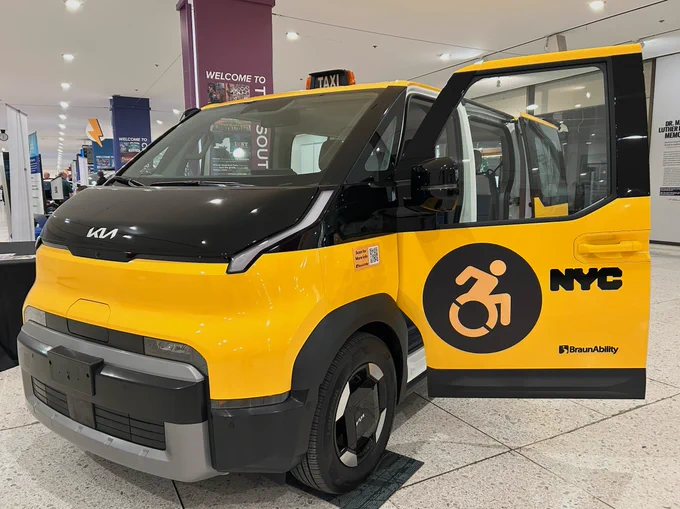 A bright yellow and black Kia electric van styled as a New York City taxi. The passenger-side sliding door is open, revealing an interior equipped for accessibility. A large circular "Wheelchair Accessible" icon and "NYC" lettering are displayed on the side of the vehicle.