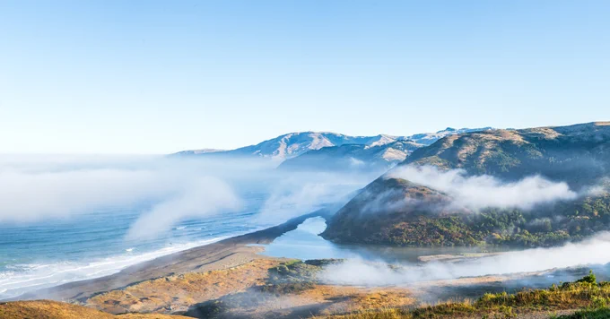 Fog rolling over a coastal mountain range with a river flowing into the ocean under a clear blue sky.