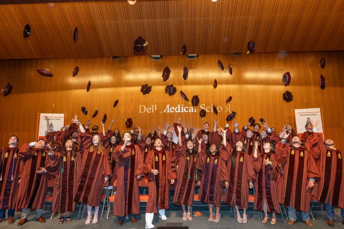 Graduates in burnt orange gowns celebrate by tossing caps in the air. The backdrop reads "Dell Medical School.