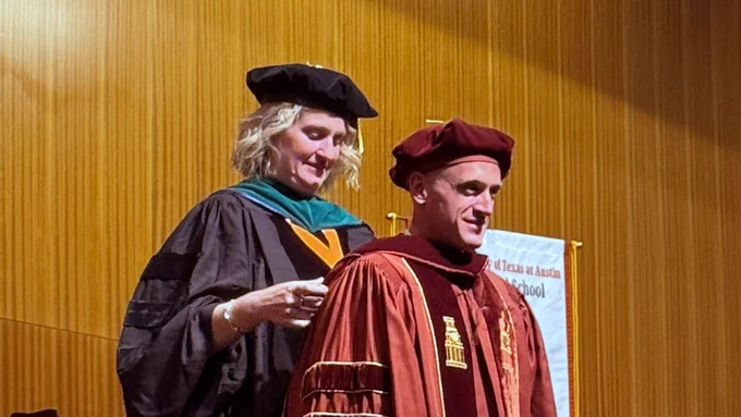 A person in graduation attire is being hooded by another individual in academic robes at a ceremony. A banner for the University of Texas at Austin is visible in the background.