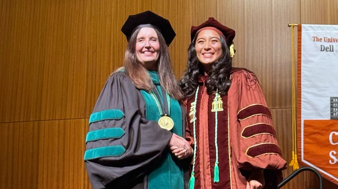 Two individuals in academic regalia stand in front of a sign showing "The University of Texas at Austin Dell Medical School." They are smiling and shaking hands.