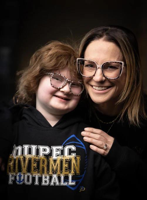 Two people are smiling at the camera. One is wearing a "DuPec Rivermen Football" hoodie. Both are wearing glasses with patterned frames.