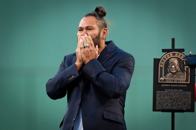 Johnny Damon stands next to his Red Sox Hall of Fame plaque with hands over his mouth looking out at Fenway Park from the infield while wearing Red Sox Hall of Fame suit coat.