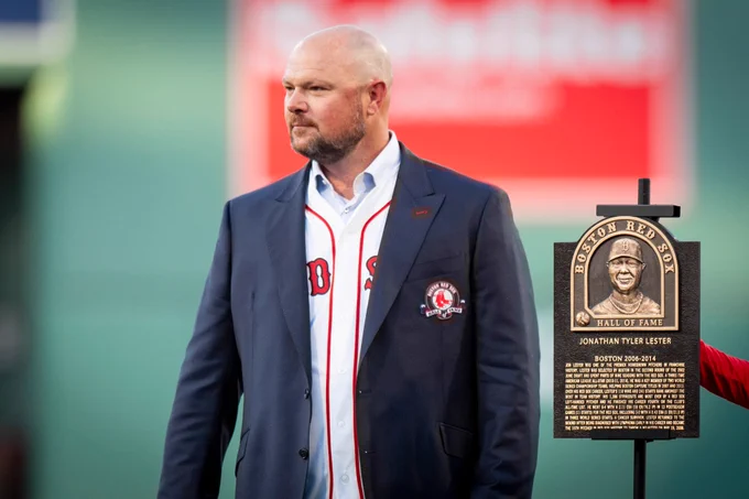 Jon Lester stands next to his Red Sox Hall of Fame plaque looking out at Fenway Park from the infield while wearing Red Sox Hall of Fame suit coat over Red Sox jersey. 