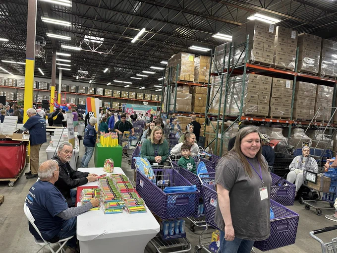A large warehouse scene with people participating in a community event. Tables are set up with school supplies like crayons and pencils. Filled shopping carts are lined up, and shelves stacked with boxes are visible in the background. The atmosphere appears busy and collaborative.
