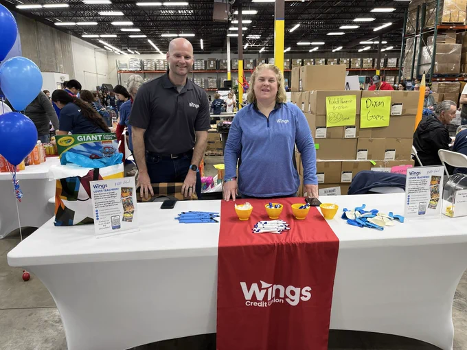 A booth at a Wings Credit Union event is set up in a large warehouse space. The table is covered with a white cloth and a red runner displaying the Wings Credit Union logo. Various promotional items and informational pamphlets are neatly arranged on the table. Blue balloons are tied to one end of the table, and there are people in the background engaged in activities.