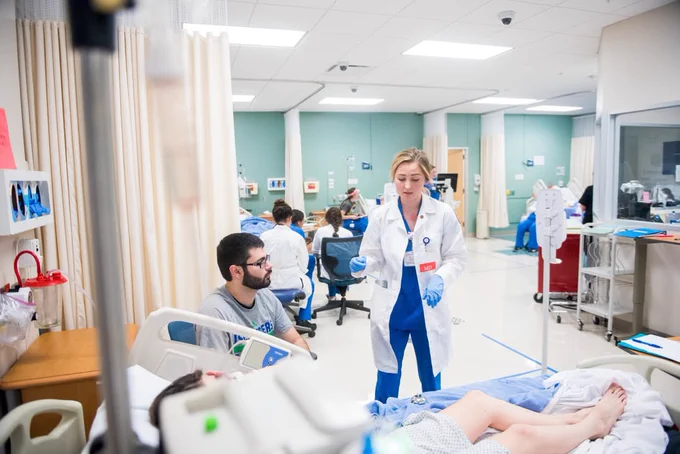 Hospital room with multiple patients and TAMU-CC student medical staff in blue and white uniforms. Staff attend to patients, with equipment and privacy curtains visible throughout the room.
