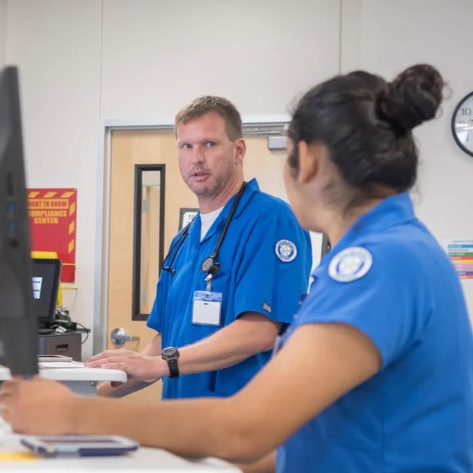 A TAMU-CC nurse instructor and student in blue medical attire, possibly healthcare professionals, conversing at a desk with screens and a clock on the wall.