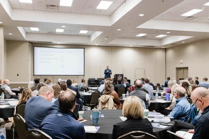 A conference room filled with attendees sitting at round tables watching a speaker near a projector screen. The presentation slide is titled "Winning Projects Today.