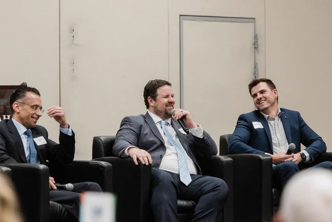 A group of individuals in suits sit on a panel, engaging in conversation and holding microphones, in a professional setting.