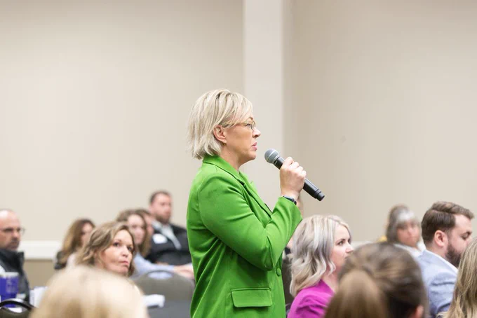 An individual in a green blazer speaks into a microphone at a conference, with seated attendees listening attentively.