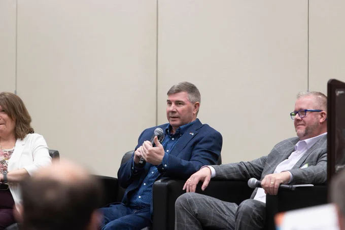 Three individuals seated and holding microphones during a panel discussion. The conversation is likely focused on a specific topic, as indicated by the formal setting and attire.
