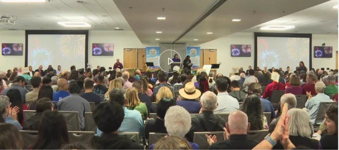 A large audience is seated in a conference hall, attentively listening to a speaker. Participants appear engaged and clapping, indicating a celebratory or supportive atmosphere.