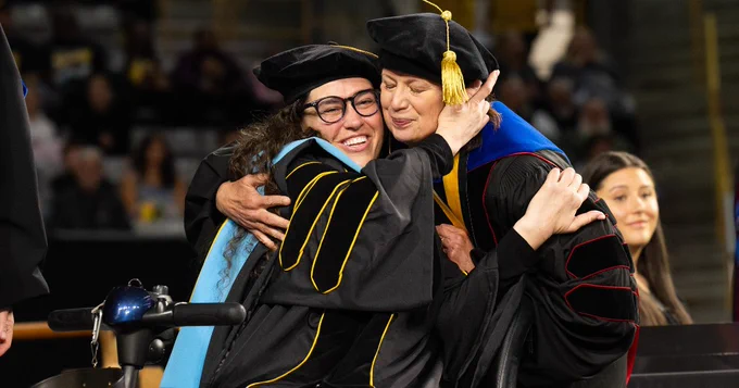 Two people in academic robes and caps embrace happily during a graduation ceremony. One wears glasses. A crowd is visible in the background.
