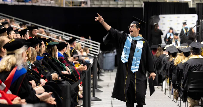 A graduate wearing a cap and gown gestures excitedly while walking down an aisle during a commencement ceremony. Seated attendees and other graduates in caps and gowns are visible in the background. 