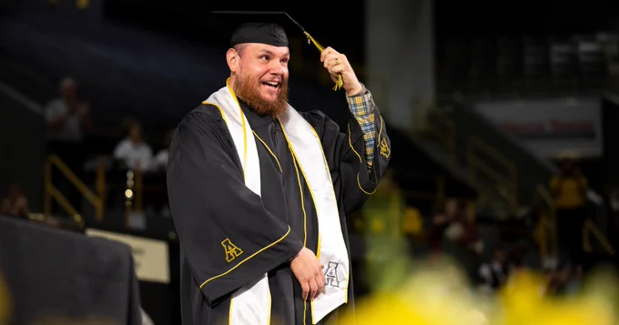 A person in a graduation cap and gown stands joyfully at a graduation ceremony. The gown features the letter "A" on the sleeve. They are holding the tassel on their cap with a lively expression. The background shows a blurred audience and a visible sign in the venue.