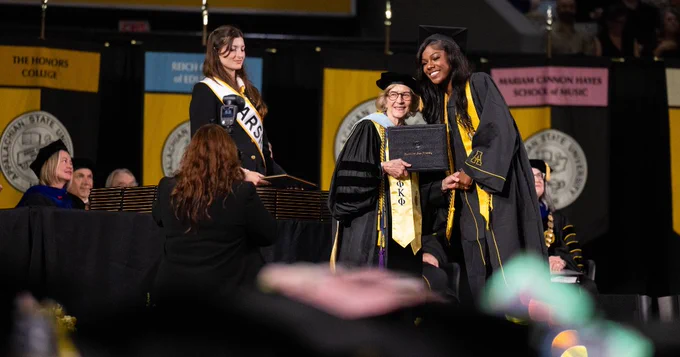 A person in academic regalia receives a diploma on stage during a graduation ceremony. Banners for various colleges are visible in the background, and others are seated and watching the event.