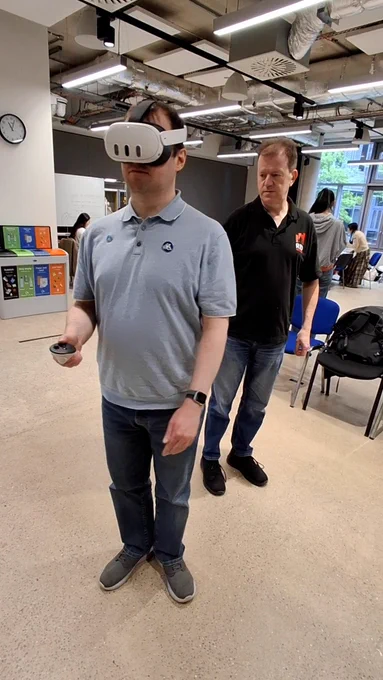 A user in a blue polo shirt navigates a room using a Meta Quest 3 headset and a hand controller. A team member from Red Nought Limited stands behind him, supervising the trial at a brightly lit indoor symposium.