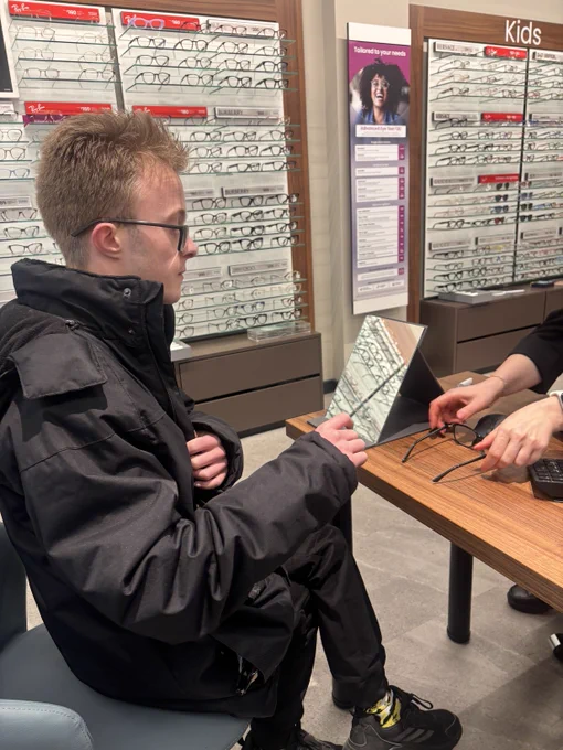 Supported Intern sitting down in a Opticain store. Rows of glasses are behind him.
