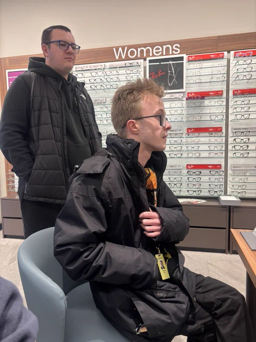 A Supported Intern wearing a bulky black coat looks to the side wearing glasses. A peer stands behind him.