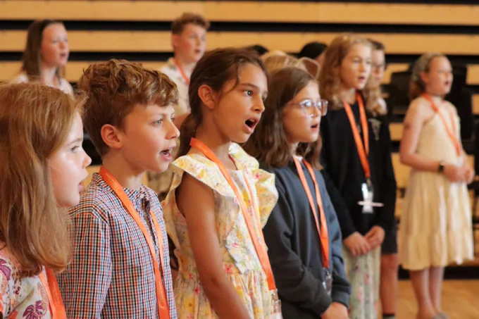 A group of children standing in a row indoors, while singing, each wearing orange lanyards and dressed in casual to semi-formal attire.
