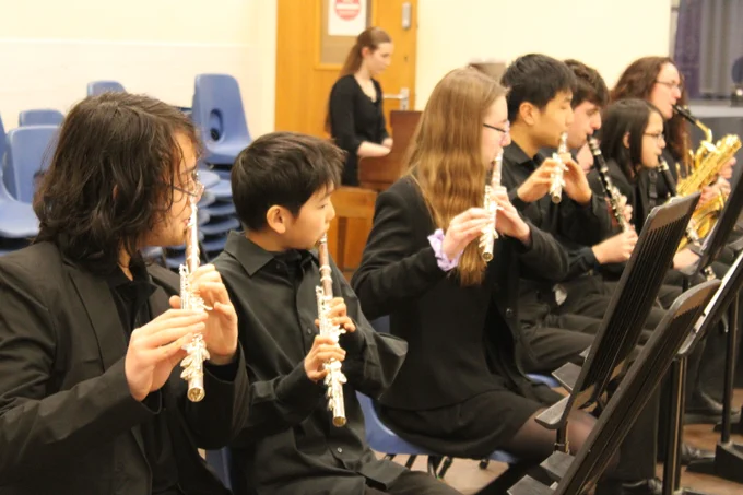 A group of young musicians seated in a row playing woodwind instruments during a rehearsal or concert indoors.