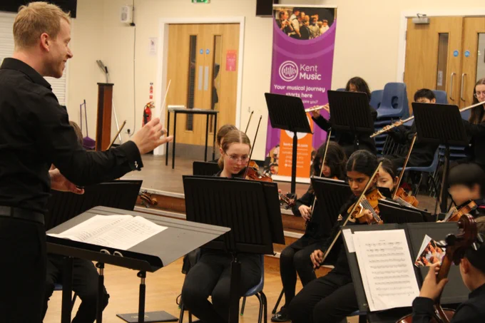 Conductor leading a youth orchestra rehearsal in a music room with musicians playing violins and sheet music stands.