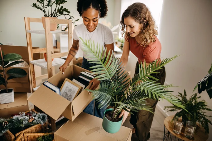 Two women unpacking a box filled with books and framed pictures in a bright room with plants around.