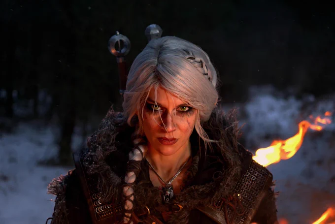 Cosplayer dressed as Ciri from The Witcher stares down the camera. Her thick silver wig is braided down one shoulder with loose strands falling across her face. Her green eyes are smudged with black makeup and black veins that extend down her cheeks. She wears thick fur-lined armor and has two swords strapped to her back.