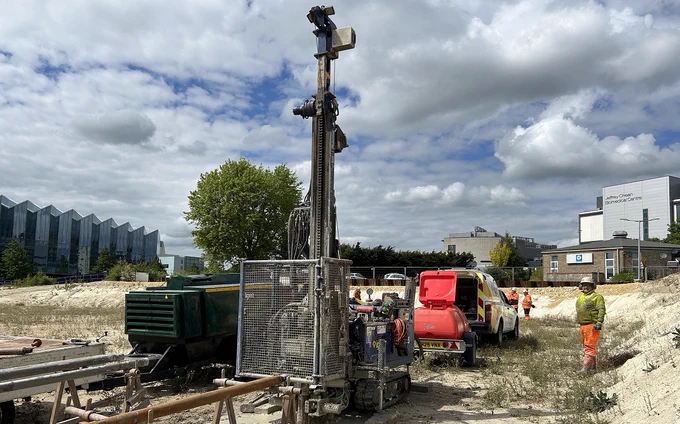 Construction site with a drilling rig, van, workers, and buildings under a cloudy sky.