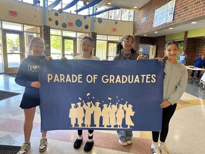 Four smiling young girls stand in the bright, modern lobby of a school, holding a large blue banner that reads "PARADE OF GRADUATES" in white capital letters. Below the text is a white silhouette of graduates in caps and gowns throwing their caps into the air.

The girls are dressed in casual school attire:

The girl on the far left wears a blue hoodie and a dark skirt.

The second girl wears a white shirt and black leggings.

The third girl wears a grey t-shirt and a purple headband.

The girl on the far right wears a light blue zip-up hoodie and black leggings.

The school's interior features brick walls, polished floors with geometric patterns, and colorful art projects hanging from the ceiling. A banner in the background mentions "Hamilton" and "Brookwood Pride."