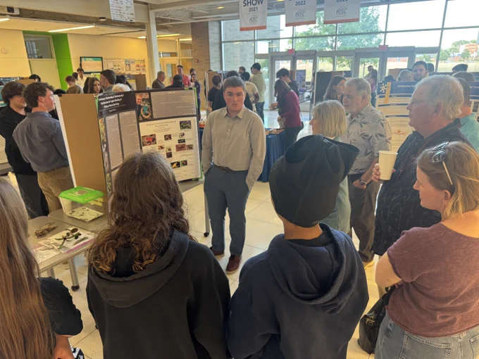 A group of people gathered around tables in the CAPS atrium, looking at science posters.