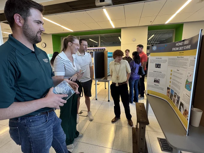 A group of parents, mentors, and friends in at CAPS atriumm, some standing near science poster displays, others engaged in discussion.