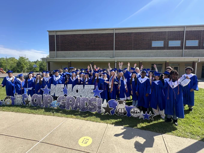 A large group of high school graduates in vibrant blue caps and gowns pose together outdoors on a sunny day. They are gathered behind a decorative yard sign that reads "WE LOVE OUR GRADS!" in glittery silver and purple lettering.

Key details of the image include:

The Graduates: Dozens of students are smiling, with several waving or raising their hands in celebration. Their gowns feature white stoles with a blue "H" logo.

The Signage: The display is embellished with various graduation-themed cutouts, including stars, a graduation cap, a "Best Grad Ever" heart, and a cartoon blue jay.

The Setting: The group is standing on a grassy area in front of a two-story brick school building under a clear blue sky. A concrete walkway is visible in the foreground.
