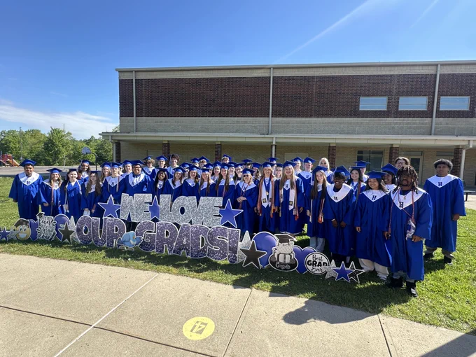 A large group of high school graduates in royal blue caps and gowns stands together on a sunny lawn for a group portrait. They are arranged behind a festive yard sign display that reads "WE LOVE OUR GRADS!" in shimmering silver and purple block letters.

Additional elements in the scene include:

The Students: The graduates are smiling and looking toward the camera; many wear white stoles featuring a blue "H" emblem.

Decorative Graphics: The sign is surrounded by graduation-themed cutouts, including stars, a blue bird mascot, a "Best Grad Ever" heart, and a tiered graduation cake graphic.

The Backdrop: The group is positioned in front of a long, two-story brick and tan school building under a bright, clear blue sky.

The Foreground: A concrete sidewalk with a yellow circular "17" marker runs across the bottom of the frame.