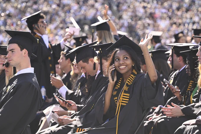A graduate wearing a cap and gown smiles and waves at FirstBank Stadium during Commencement. 