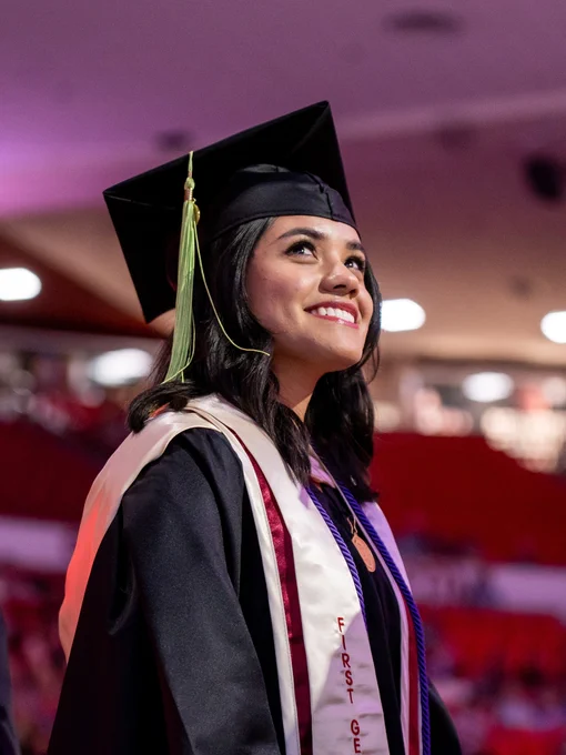 A smiling graduate in a black cap and gown looks upward during a commencement ceremony, wearing a "First Generation" sash, a purple honor cord, and a green tassel, with a red-seated arena visible in the background.