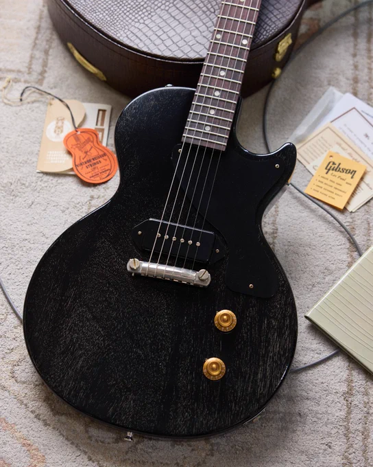 A Gibson Custom 1957 Les Paul Junior Reissue electric guitar in Silver Fox finish lies on a textured rug. Nearby are a guitar case, a Gibson instruction booklet, tags, and other paperwork.