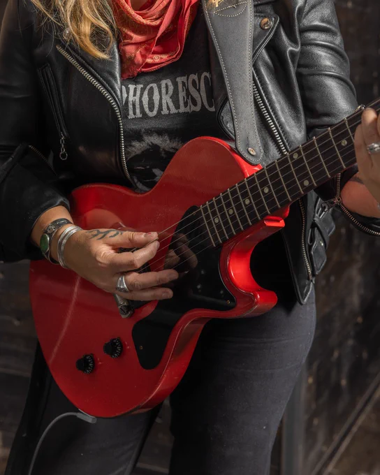 A person playing a bright Cardinal Red Gibson Custom 1957 Les Paul Junior Single Cut Reissue electric guitar, wearing a leather jacket and a red scarf. Only the upper body and hands are visible.