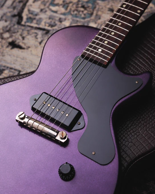 A close-up of a Purple Sparkle Gibson Custom 1957 Les Paul Junior Reissue electric guitar resting on a textured surface. The guitar features a black pickguard, six strings, and various control knobs.