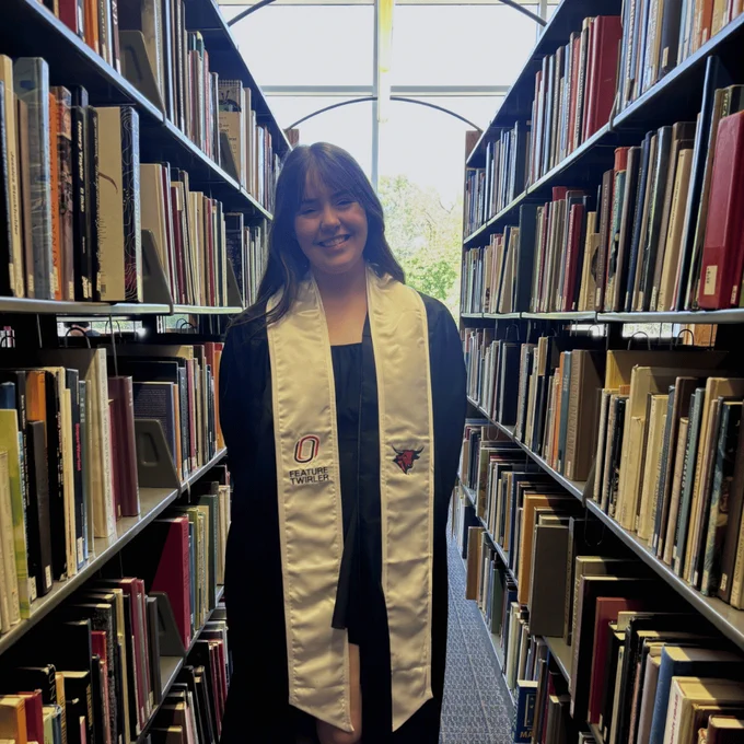 A young woman standing in between two library stacks. She is wearing a graduation gown and sash and smiling at the camera.