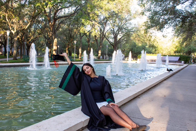 Sara Arroyave Soto in a graduation gown sits by a fountain, holding a cap in the air. The background features trees and multiple water jets, suggesting a celebratory scene in a peaceful outdoor setting.